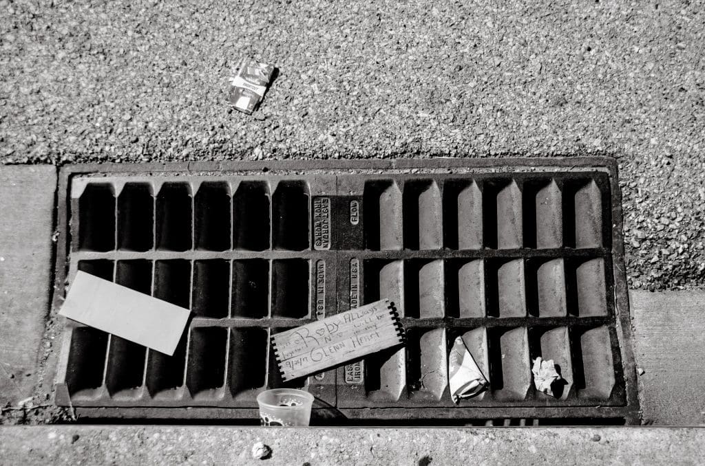 a street drain with trash and a wooden board with the text "Roby N Glenn always will be in my heart" written on it