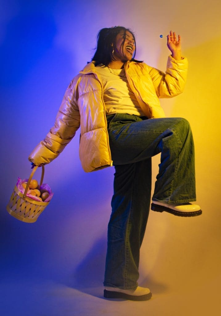 a girl holding a basket of lemons, wearing a yellow jacket and blue jeans in front of a blue and yellow background