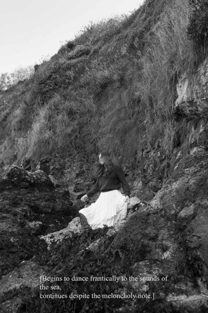 a girl in a dark long sleeve shirt and white skirt sitting on a rocky hill with the text "Begins to dance to the sounds of the sea, continues despite the melancholy note" written in white on the bottom half of the image