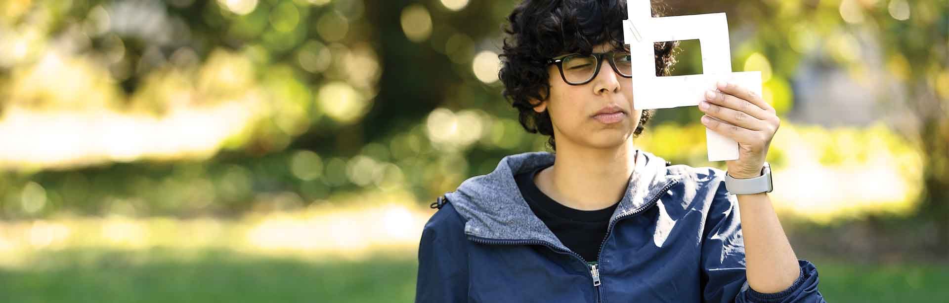 student sitting in a field looking through a drawing square