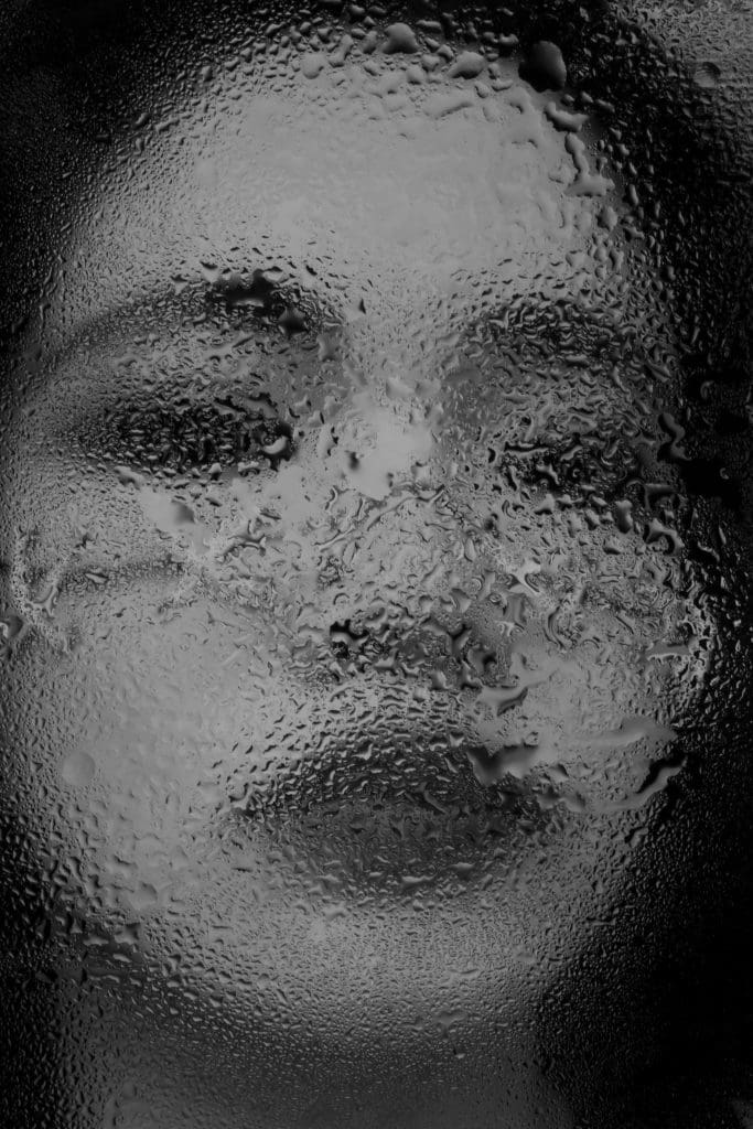 a black and white image of a girls face behind a blurred water dripped glass surface