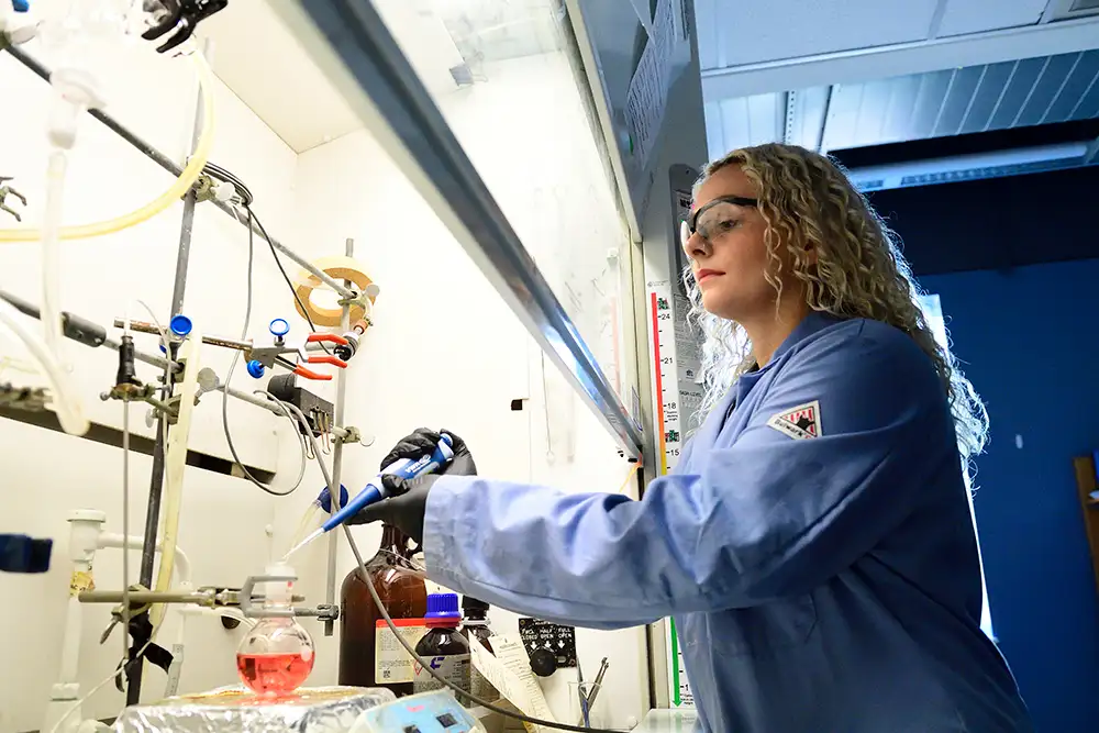 A member of Thomas Kempa's lab pipetting liquid into a glass container under a chemistry fume hood.