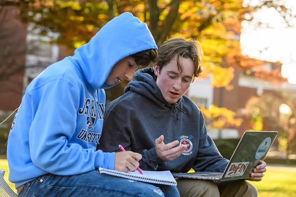 two students outdoors on campus - one talking and the other taking notes