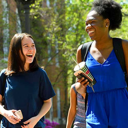Two students smiling as they walk through campus on a sunny summer day.