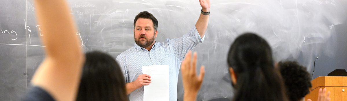 a faculty member raising his hand in front of a chalkboard