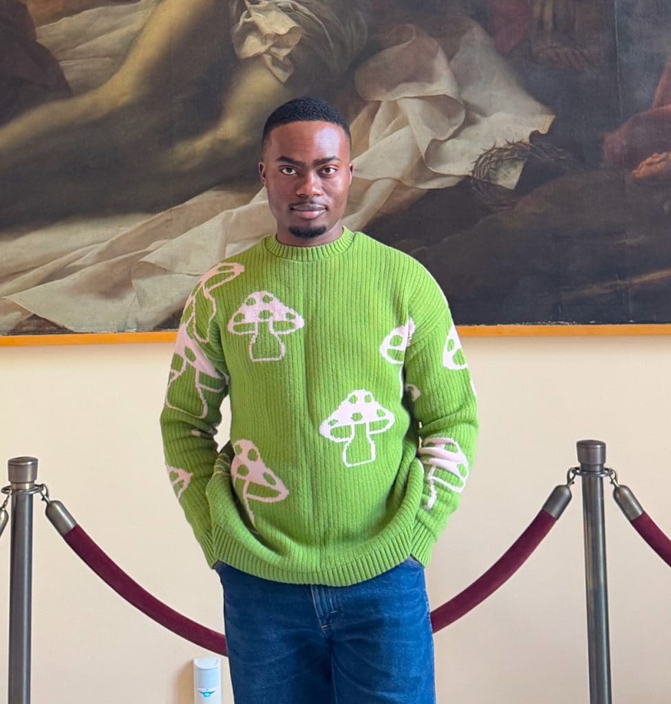 Writing Seminars MFA alumni Olakunle Ologunro stands in front of a large oil painting in a museum, wearing a bright green sweater.